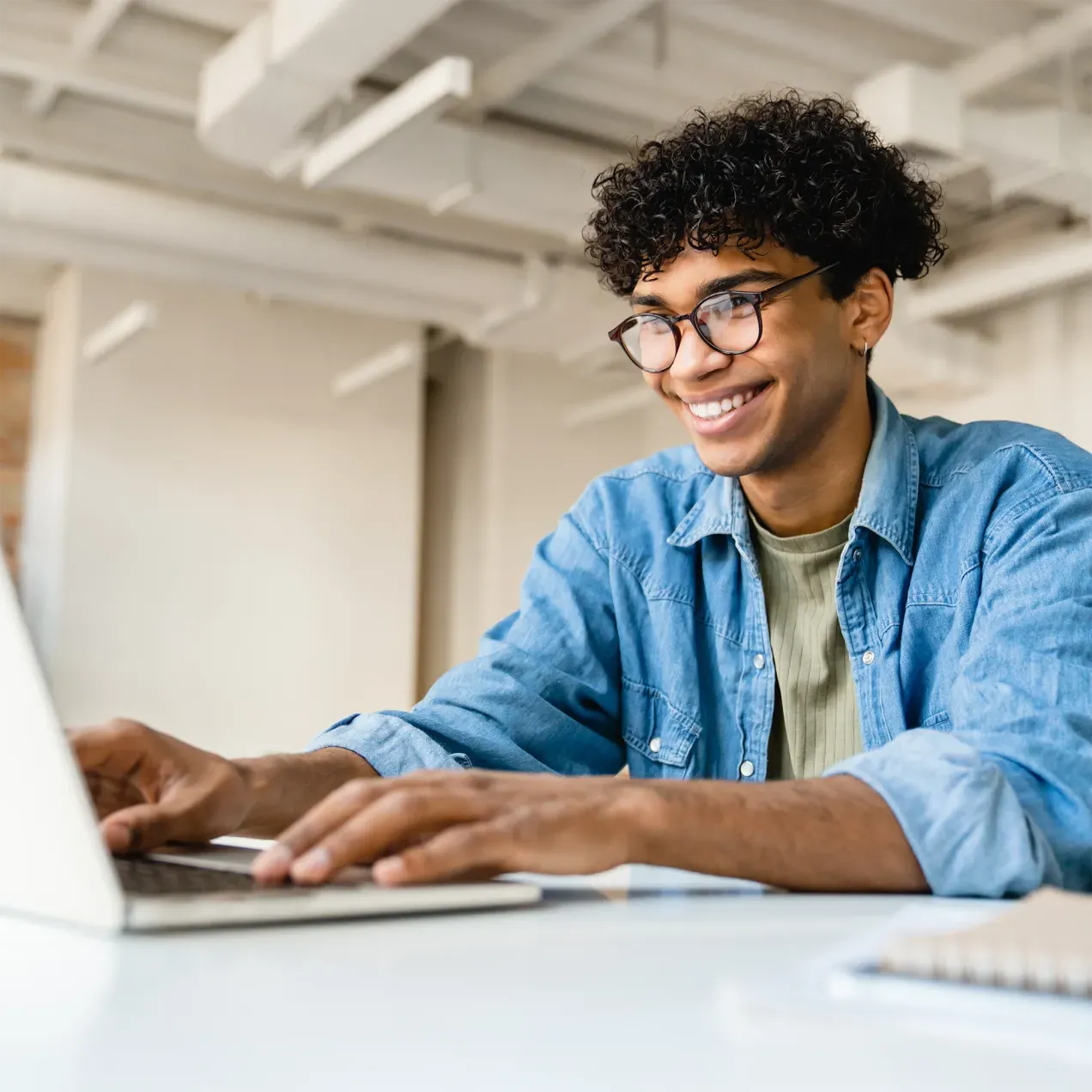 A Herzing University information technology student in a denim shirt and glasses smiles while working on a laptop in a modern, well-lit workspace.